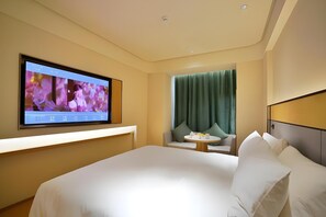Une chambre d’hôtel avec un grand lit, un téléviseur fixé au mur, une petite table ronde avec une chaise, et une fenêtre avec des rideaux verts.