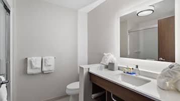 Un baño moderno con lavamanos, espejo y retrete blancos. Hay toallas colgadas en la pared y se ve una ducha en el reflejo.