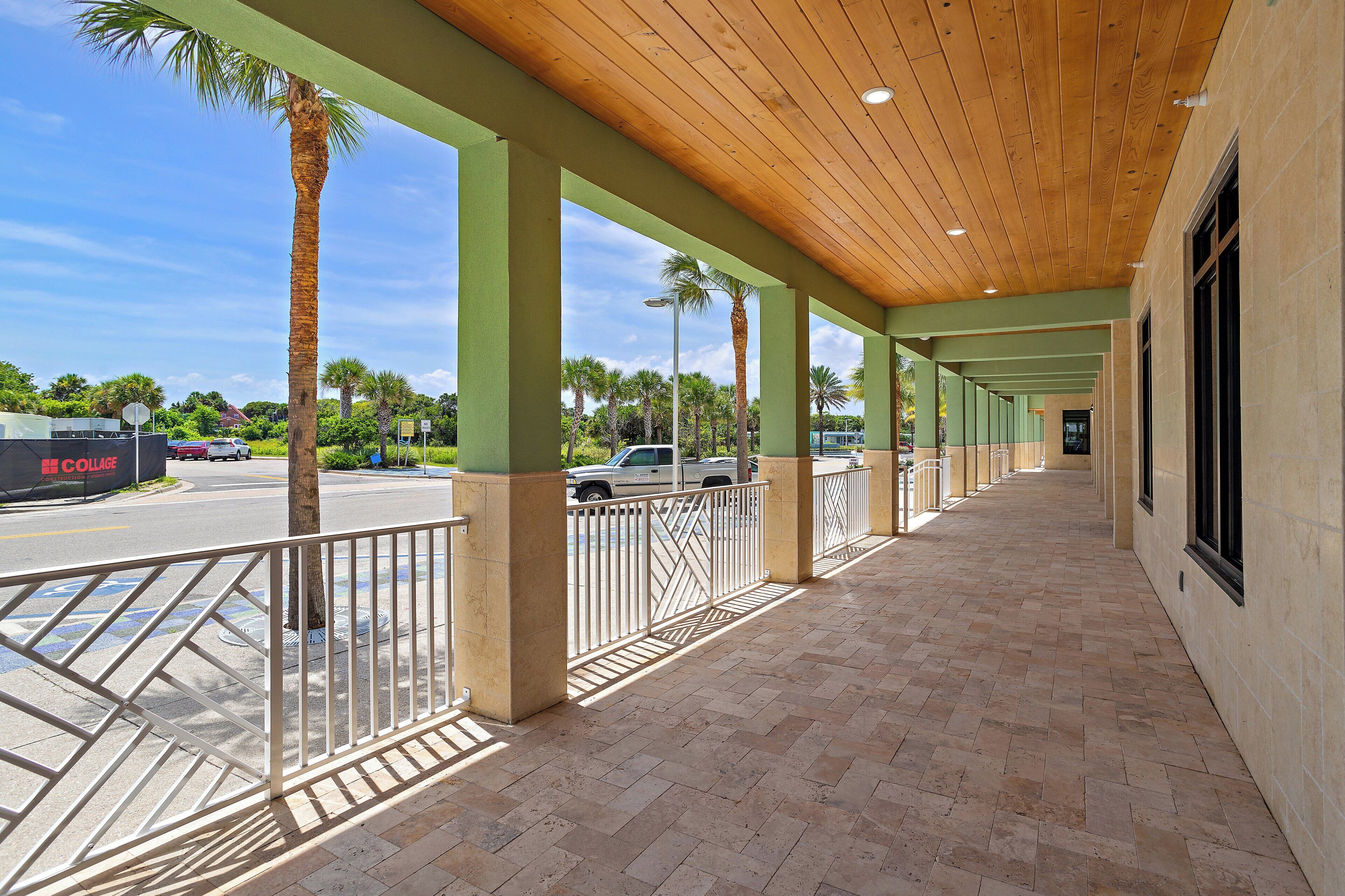 A covered walkway with a tiled floor, palm trees, and a view of parked cars and a building in the background.