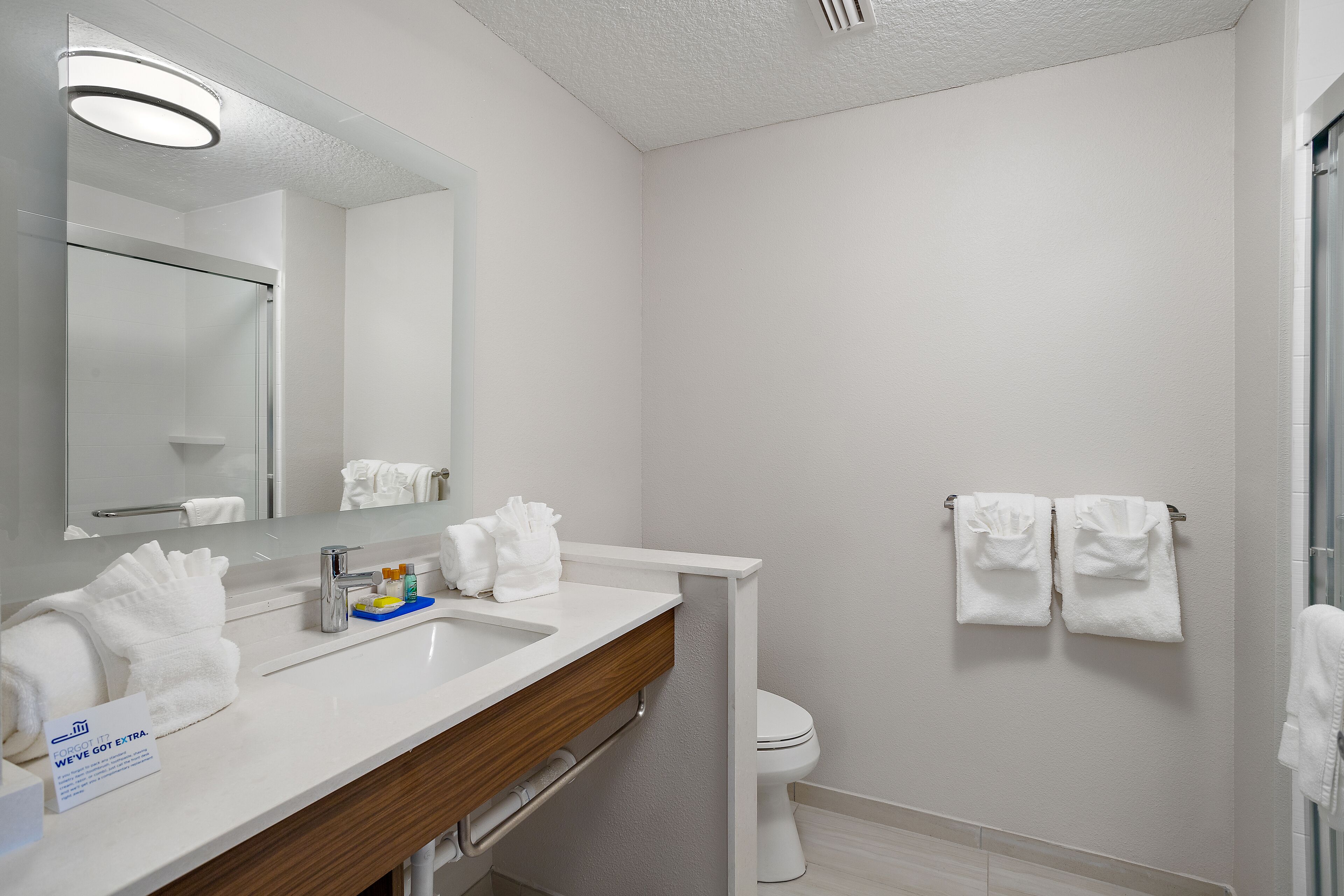 Un baño moderno con lavamanos, espejo y inodoro blancos. Hay toallas en la pared y un cartel que indica que hay toallas adicionales disponibles.