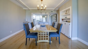 A dining room with a wooden table, blue upholstered chairs, a chandelier, and a kitchen in the background.