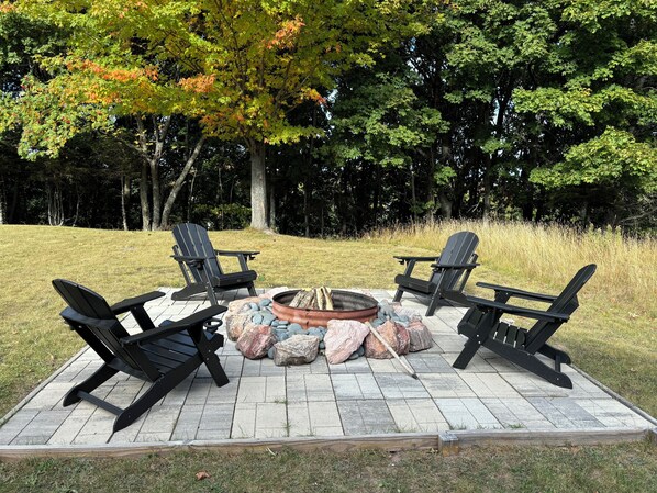 A circular fire pit with four Adirondack chairs around it, set on a paved patio with a grassy lawn and trees in the background.