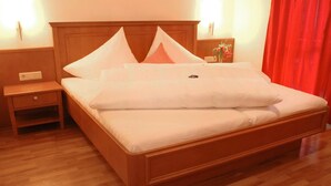 Un lit bien fait, avec du linge de lit blanc et des oreillers, une table de chevet en bois avec un tiroir, et un rideau rouge en arrière-plan.