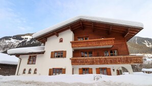 Un chalet en bois à deux étages, avec un toit et un balcon couverts de neige, situé au pied d’une chaîne de montagnes.