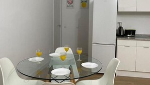 A compact kitchen with white cabinets and a refrigerator, a glass dining table with four chairs, and two wall decorations.