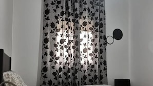 A bedroom with a bed, a bedside table, a lamp, a wall-mounted light, and a curtain with a leaf pattern.