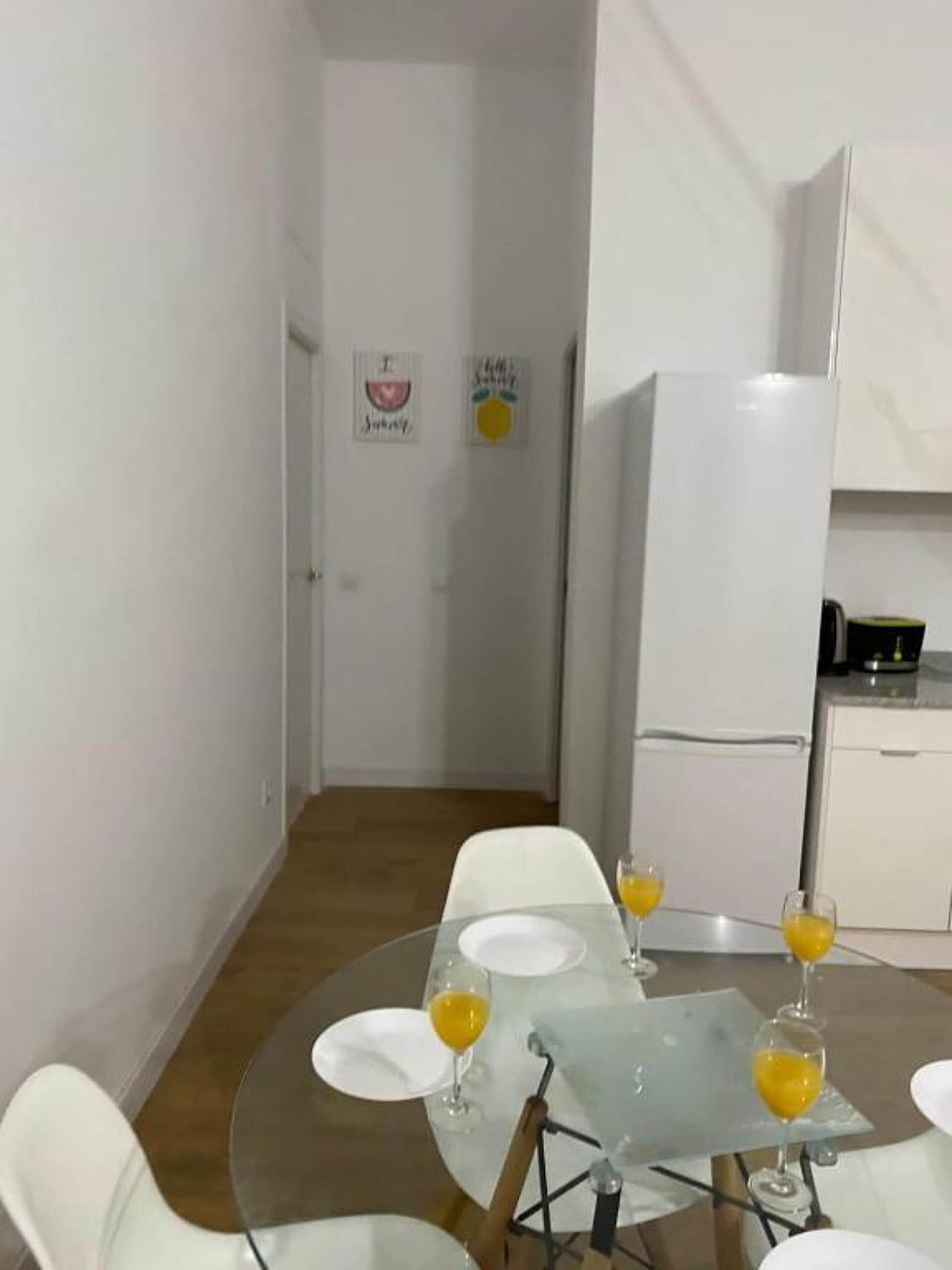 Comedor con mesa de vidrio, sillas blancas y un refrigerador al fondo.