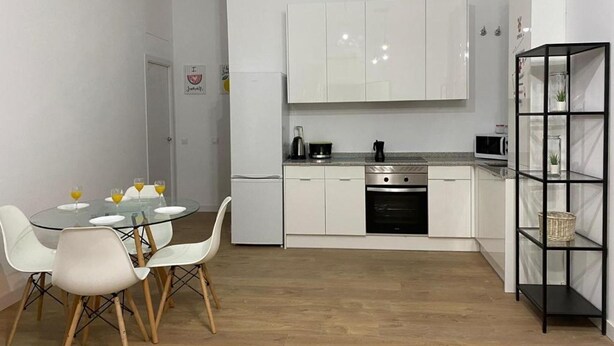 A compact kitchen with white cabinets, a dining table with chairs, a black metal shelf, and a microwave.