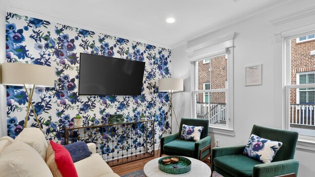 Un salon avec un papier peint à motifs floraux, un téléviseur à écran plat, deux fauteuils verts, un canapé couleur crème et une table basse agrémentée d’un vase décoratif.