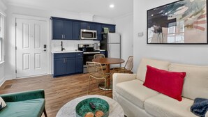 Une cuisine moderne avec des meubles de couleur bleu foncé, un réfrigérateur blanc et un four à micro-ondes. On y trouve un coin repas avec une table ronde et deux chaises en osier. Dans le salon, il y a un canapé de couleur crème agrémenté d'un coussin rouge.