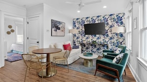 Un salon moderne avec une table ronde en bois, deux chaises en fil de fer doré, un canapé blanc, un fauteuil turquoise et un tapis à motifs.