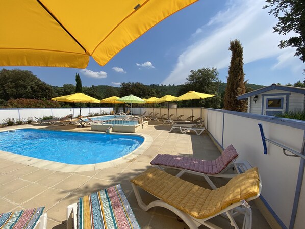 Area piscina con diverse sdraio e ombrelloni, circondata da un muro bianco e vista su alberi e colline.