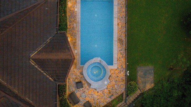 Eine Vogelperspektive auf einen rechteckigen Swimmingpool mit Steinbord, umgeben von einer Steinterrasse mit Sitzgelegenheiten und einem kleinen Brunnen.