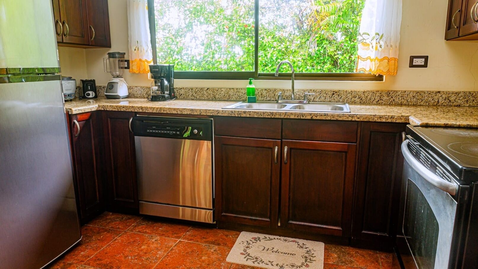 A kitchen with dark wooden cabinets, stainless steel appliances, and a window with a view of greenery.