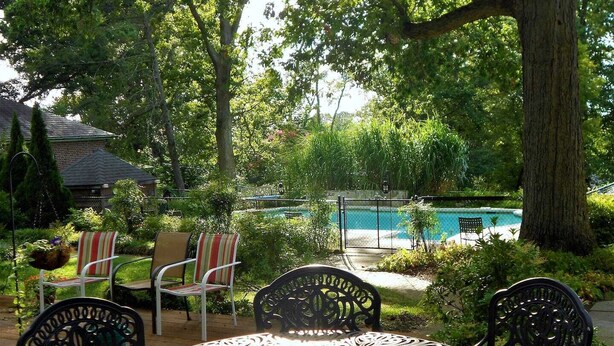 An outdoor patio with a metal table and chairs, a swimming pool in the background, and lush greenery. - Rollingside Guest Suite (Catonsville)
