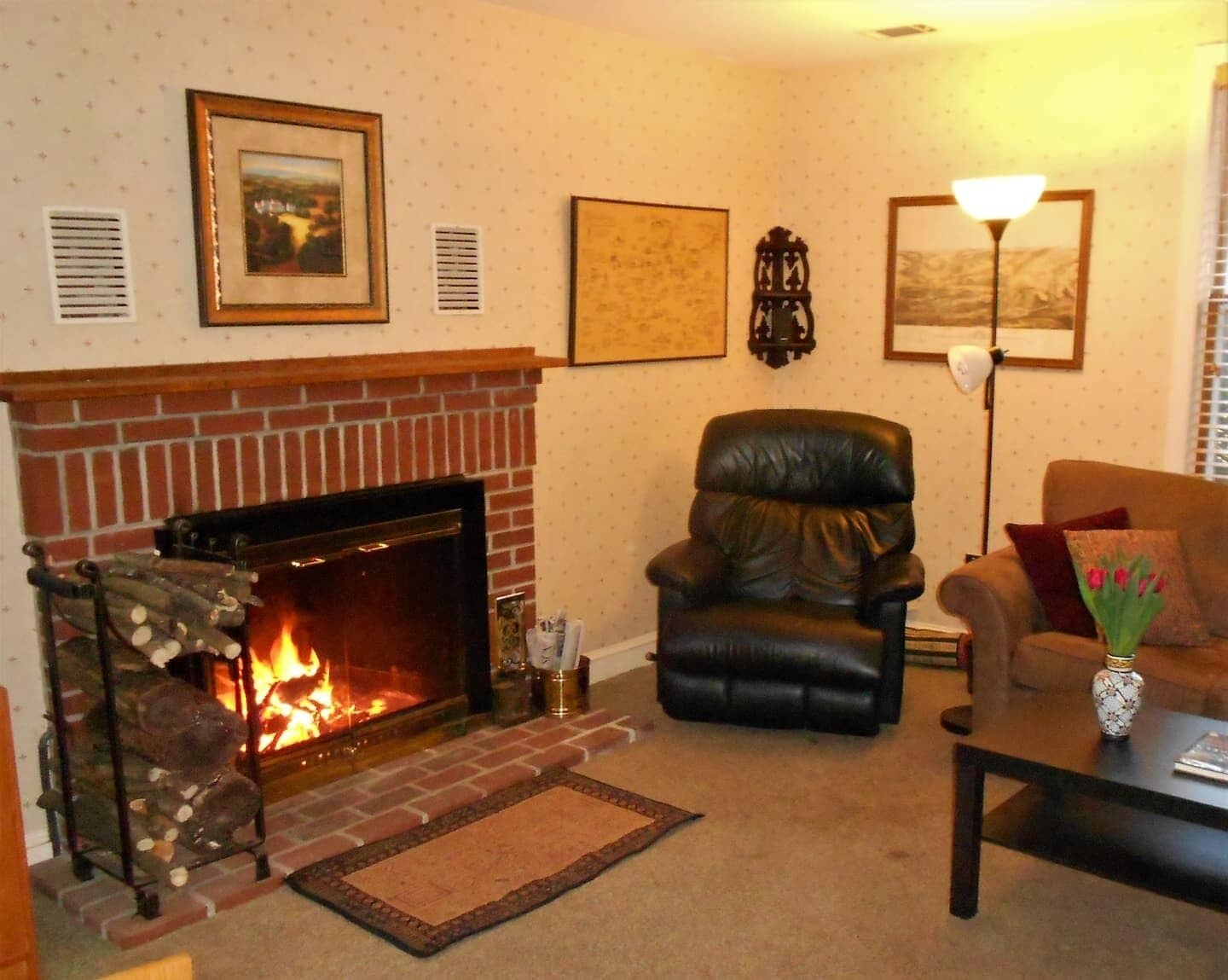 A living room with a brick fireplace, a leather armchair, a sofa, a coffee table, and wall-mounted artwork.