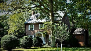 A brick house with a front porch, surrounded by well-maintained lawns and trees. - Rollingside Guest Suite (Catonsville)