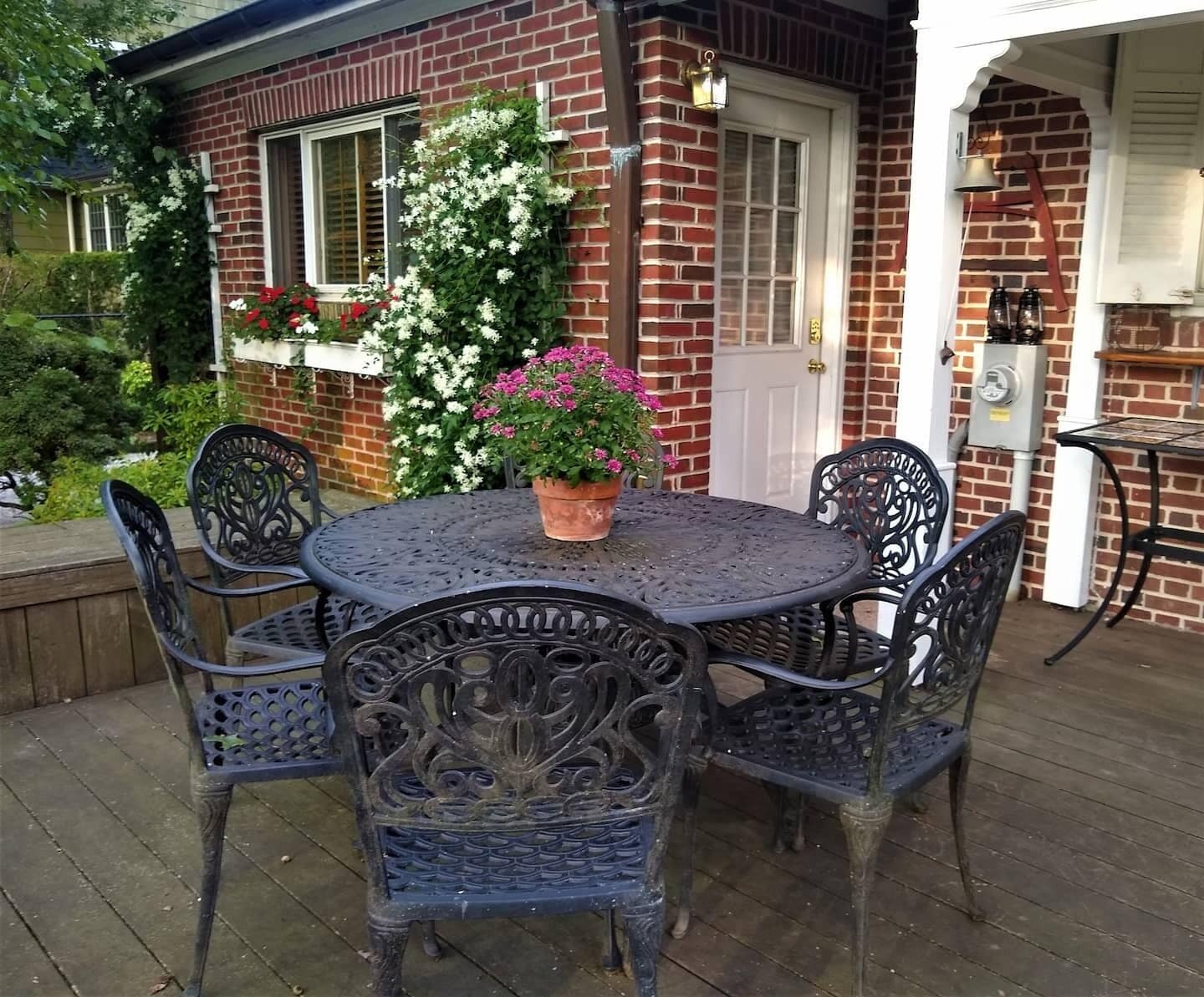 Une terrasse avec une table ronde en métal et quatre chaises, entourée d’une maison en briques avec une porte et des fenêtres blanches.