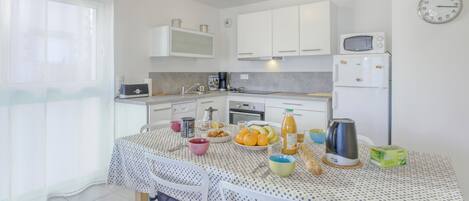 Una cocina con armarios blancos, microondas, reloj y una mesa con frutas y una tetera.