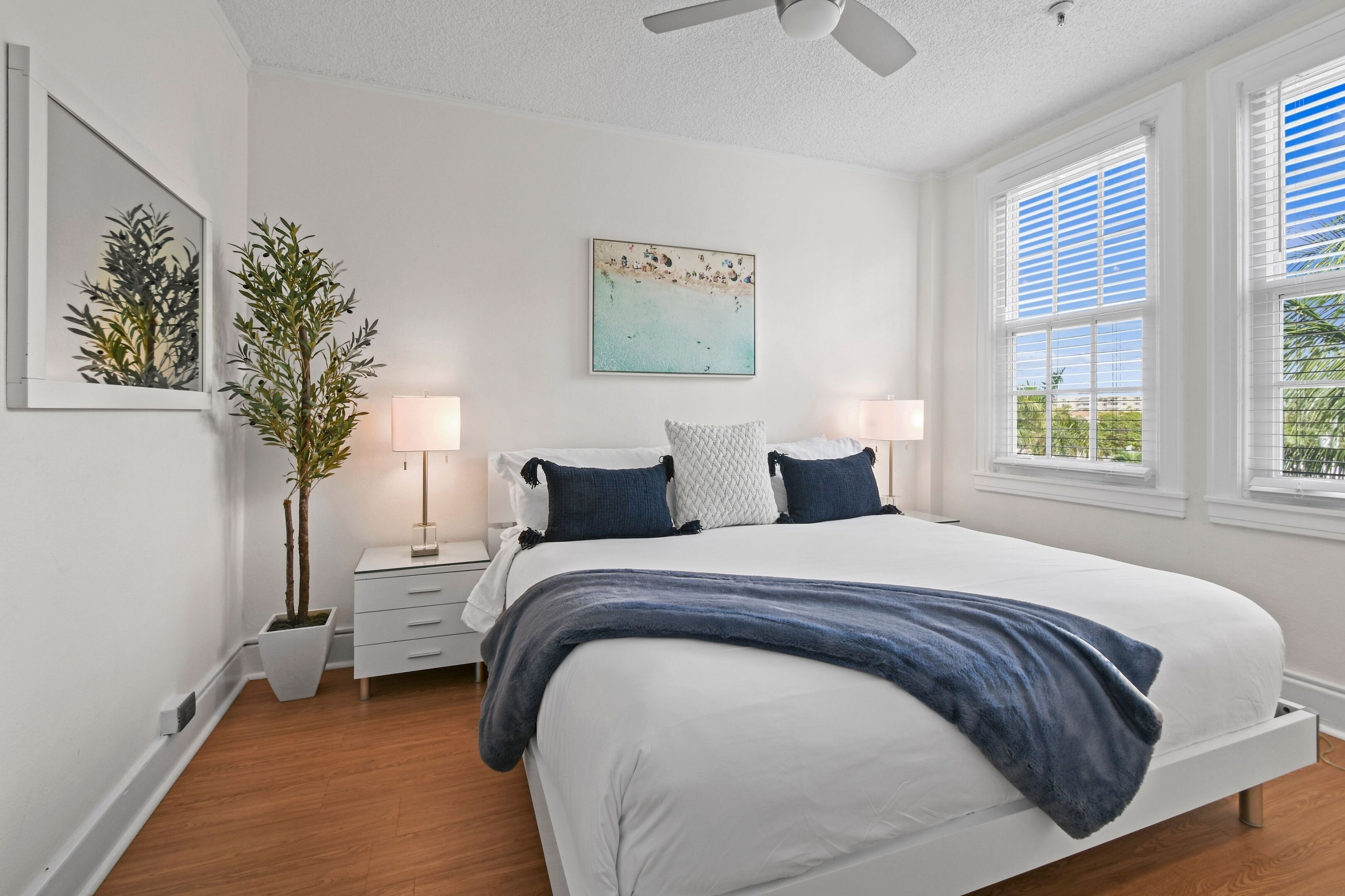 A bedroom with a bed, bedside tables, a nightstand, a potted plant, and a window with blinds.