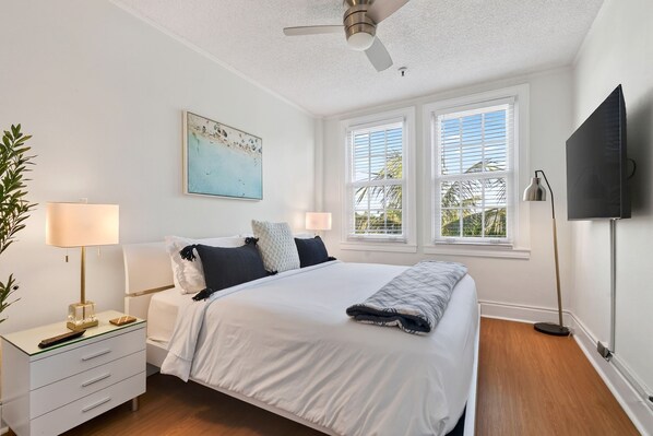 A bedroom with a bed, bedside lamps, a nightstand, a desk, a television, and a ceiling fan. - Condo/Pool/StepsToBeachDine/Island 1br (Palm Beach)