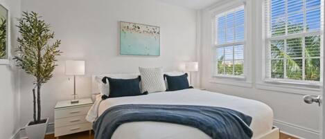 A bedroom with a bed, two bedside tables, a ceiling fan, a window with blinds, and a potted plant.