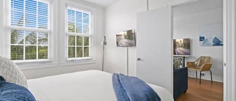 Quarto com cama, duas janelas com persianas, televisão e uma cadeira.