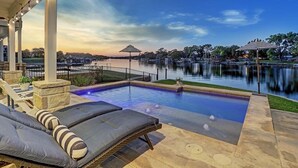Una piscina exterior con sillones de descanso y sombrillas, rodeada por un lago y un parque.