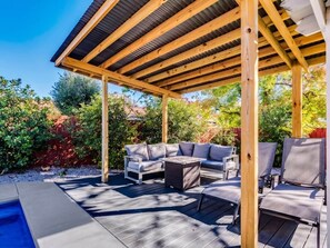 Een overdekt terras met een zwembad, een houten pergola en omliggend groen.