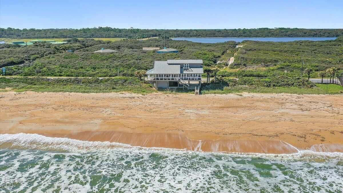 A coastal property with a large house, a sandy beach, and a body of water in the background.