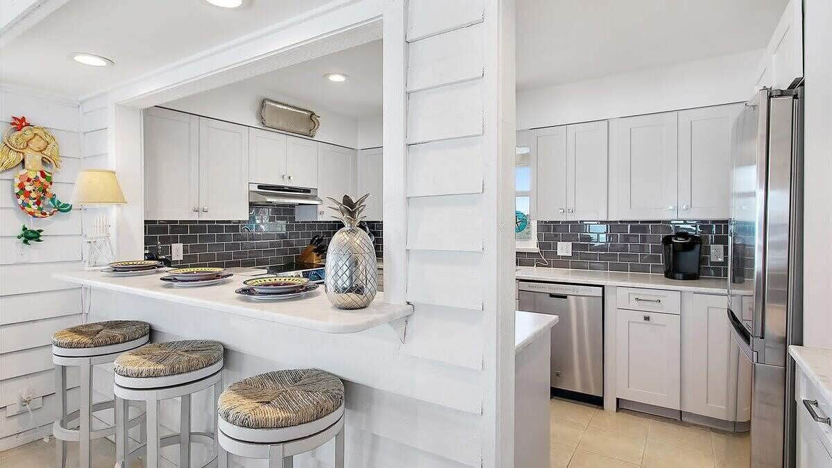Een moderne keuken met witte kastjes, een zwart betegelde spatwand en roestvrijstalen apparatuur. Er staan drie barkrukken aan het aanrecht en een decoratieve ananas als middelpunt.