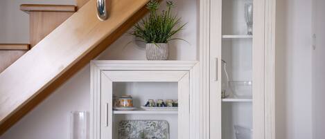 Una escalera con escalones de madera y una pared blanca. A la derecha, un armario blanco con puertas de cristal, y a la izquierda, una planta en maceta.