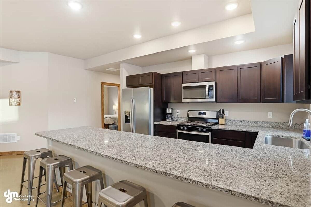 A modern kitchen with granite countertops, dark cabinetry, stainless steel appliances, and a bar area with stools.