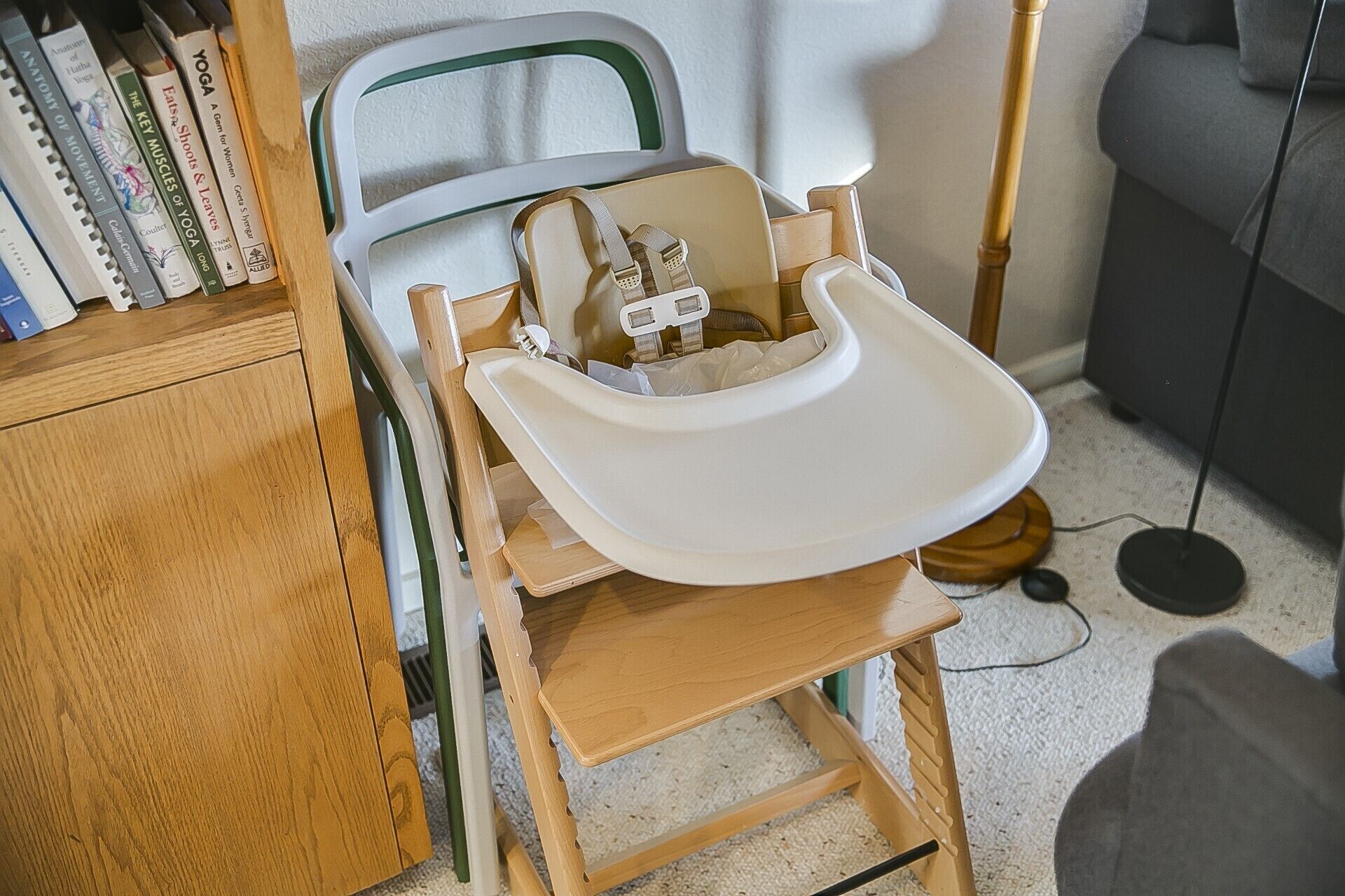 A wooden high chair with a white tray, positioned next to a bookshelf and a floor lamp.