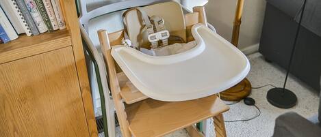 A wooden high chair with a white tray, positioned next to a bookshelf and a floor lamp.