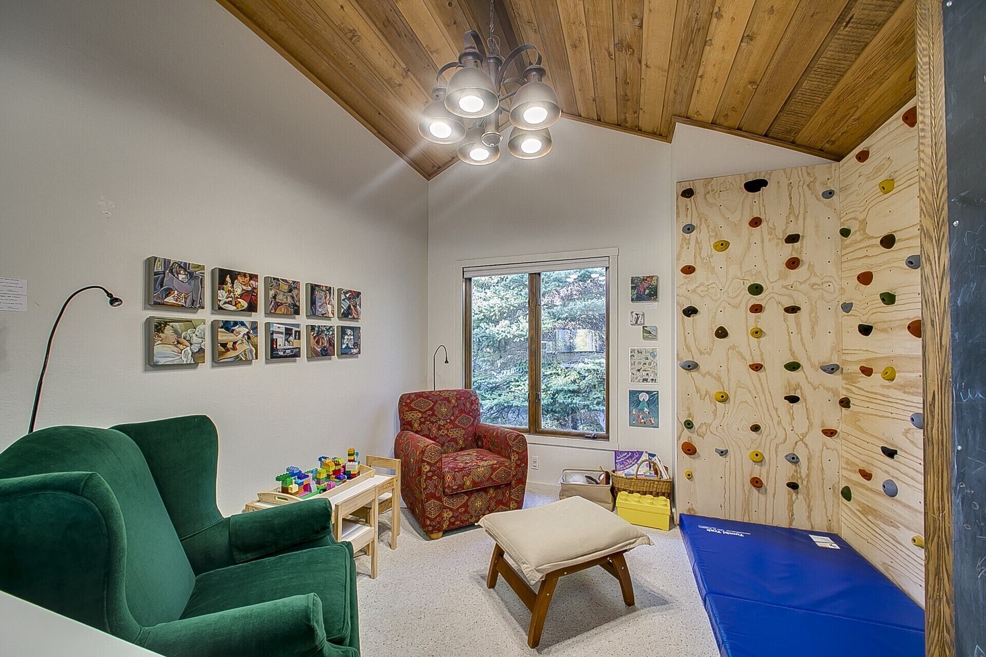 A room with a climbing wall, a green chair, a small table with toys, and a window with a view of trees.