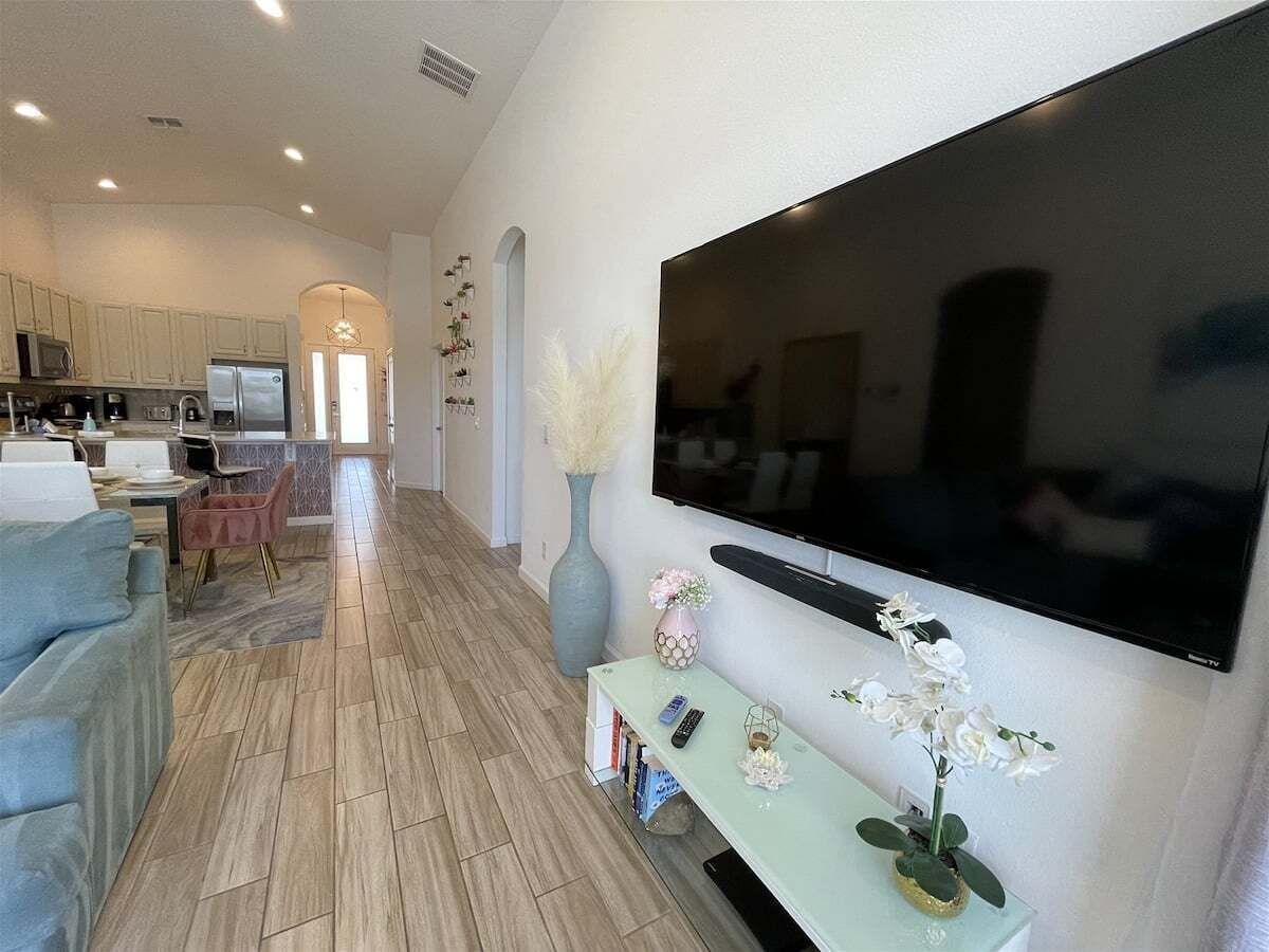 A modern living room with a flat-screen TV, a dining area with chairs, a sofa, and a kitchen in the background.