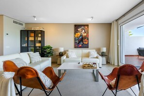 Un salon moderne avec un canapé, une table basse et des fauteuils, un tableau au mur et une porte-fenêtre coulissante donnant sur un espace extérieur.