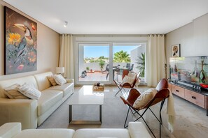 A living room with a sofa, coffee table, and chairs, a painting, a TV, and a sliding glass door leading to an outdoor patio.