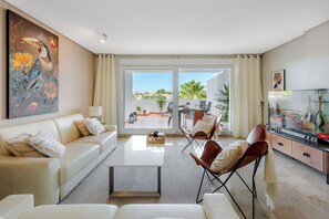 Un soggiorno moderno con un divano bianco, un tavolino da caffè e una televisione. È presente un balcone con mobili da esterno e vista sulla città.