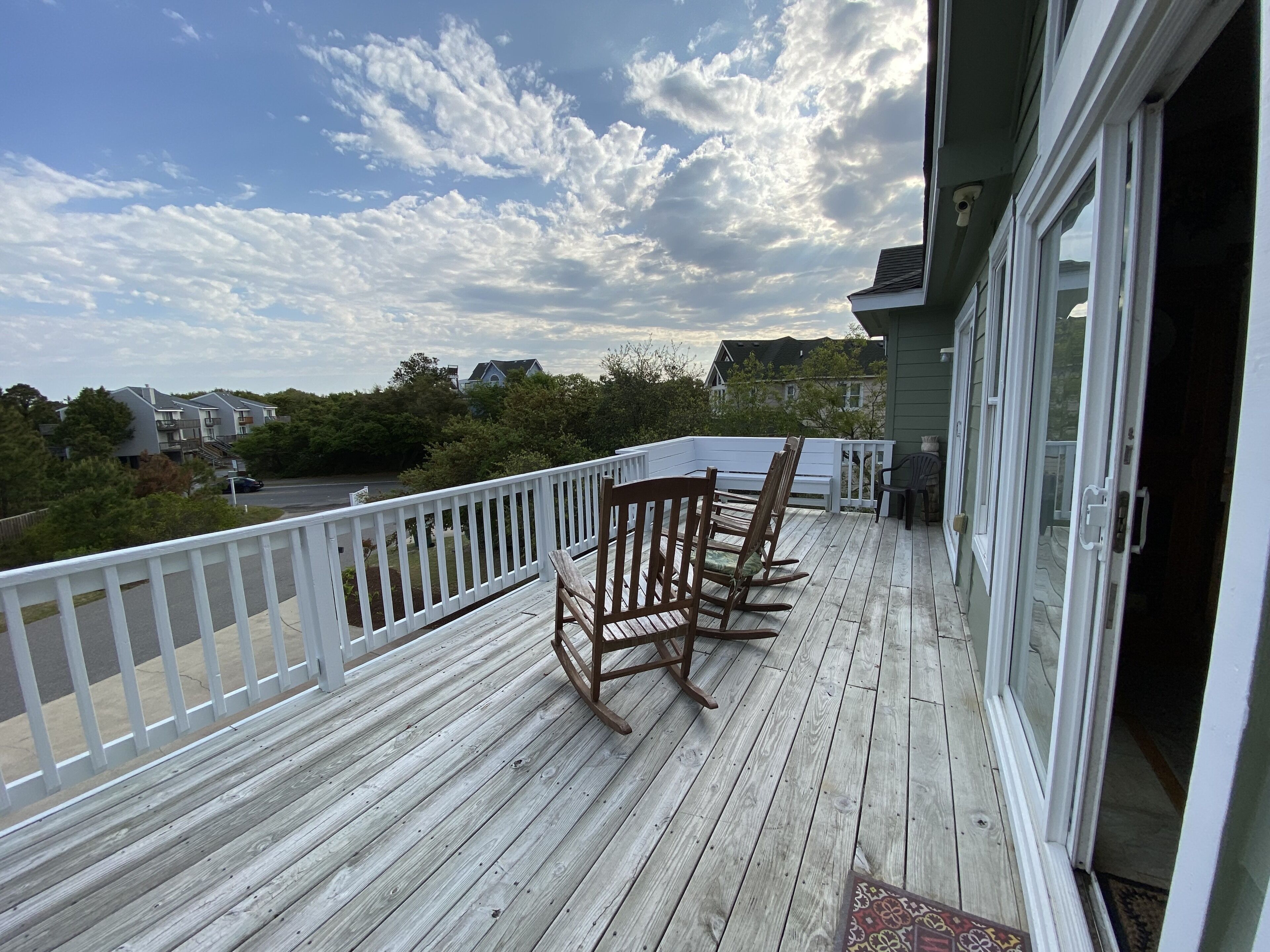 Une terrasse en bois avec des balustrades blanches, une rangée de fauteuils à bascule en bois et, au loin, une vue sur des maisons et des arbres.