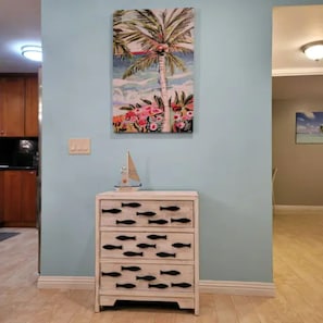 Een kamer met lichtblauwe wanden, een houten kast met vis-vormige knoppen, een klein modelzeilboot en een schilderij van een palmboom en bloemen.