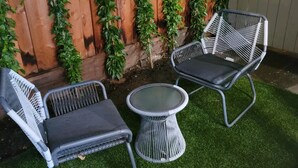 Eine Terrassenfläche mit zwei Korbstühlen und einem kleinen runden Tisch auf Kunstgras, vor einer Holzzahlung mit Kletterpflanzen.