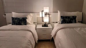 Ein Hotelzimmer mit zwei Betten, jeweils mit weißem Kopfteil und einem Kissen mit Blattmuster. Dazwischen befindet sich ein Nachttisch mit einer Lampe und einem kleinen Tisch.