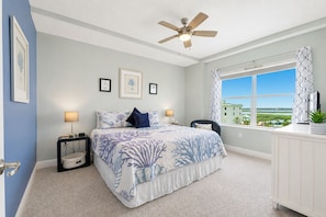 A bedroom with a bed, two bedside tables, a ceiling fan, a window with a view, and framed pictures on the wall.