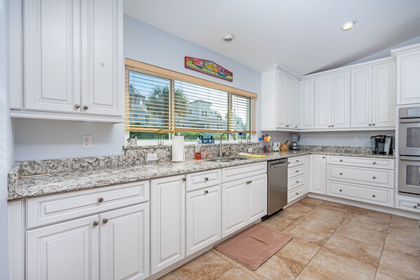 A kitchen with white cabinets, granite countertops, a stainless steel dishwasher, and a large window with blinds. - Beach-side Pool Home - Hope's Haven (Port Orange)