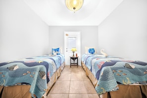 A room with two beds, each with a blue and white ocean-themed bedspread, a small table with a lamp, and a door in the background.