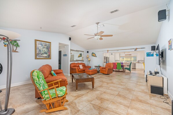 A spacious living room with a ceiling fan, orange furniture, a rocking chair, and a view into the kitchen. - Beach-side Pool Home - Hope's Haven (Port Orange)