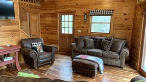 L’intérieur chaleureux d’un chalet, avec un revêtement mural en bois, un téléviseur fixé au mur, un fauteuil en cuir marron, un canapé en cuir marron et un pouf assorti.