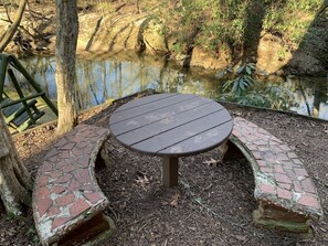 Une table ronde en bois, entourée d’un banc en briques, est installée dans un espace boisé, avec un étang en arrière-plan.